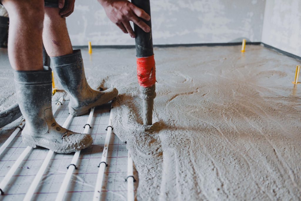 Eine Person in Gummistiefeln gießt nassen Beton über ein Gitter aus weißen Rohren, wahrscheinlich für die Fußbodenheizung, im Inneren eines im Bau befindlichen Gebäudes - ein entscheidender Schritt, bevor die Verlegung der Fliesen beginnt.