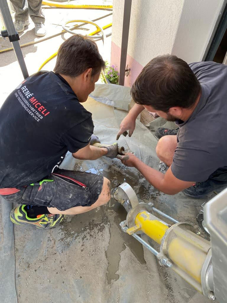 Zwei Arbeiter in Stutensee knien auf einer Plane und montieren oder reparieren Metallrohre für eine Bautrocknungsanlage, die an einer gelben Industriemaschine befestigt ist. Einer trägt schwarz, der andere grau. Werkzeuge und verschüttete Flüssigkeit sind auf dem Boden zu sehen.