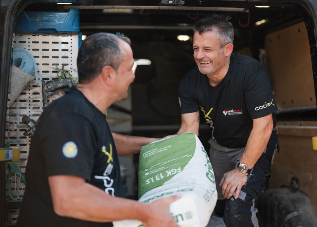 Zwei Männer in schwarzen Hemden arbeiten lächelnd zusammen, um einen schweren Sack aus dem hinteren Teil eines Lieferwagens in Stutensee auszuladen, der möglicherweise Materialien für ein Bautrocknungsprojekt transportiert. Ein Mann übergibt den Sack an den anderen im Inneren des Fahrzeugs.