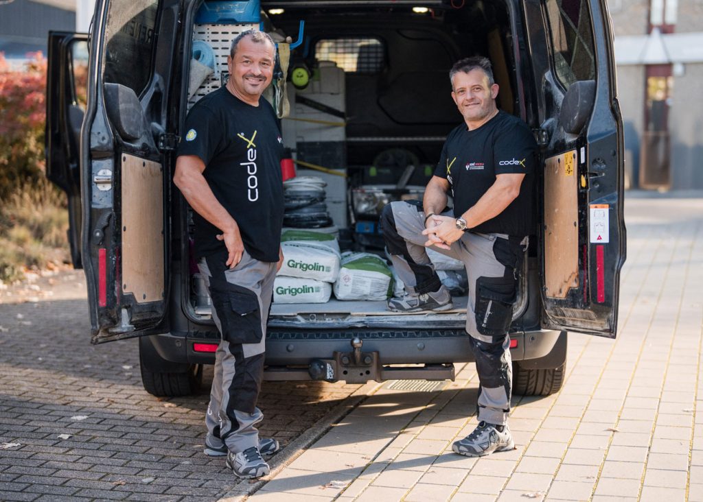 Zwei Männer in Arbeitsuniformen, wahrscheinlich Fliegenleger aus Stutensee, stehen am offenen Heck eines mit Werkzeug und Baumaterialien gefüllten Lieferwagens. Sie lächeln und posieren für das Foto auf einem gepflasterten Außengelände, möglicherweise nach einer anstrengenden Arbeit.