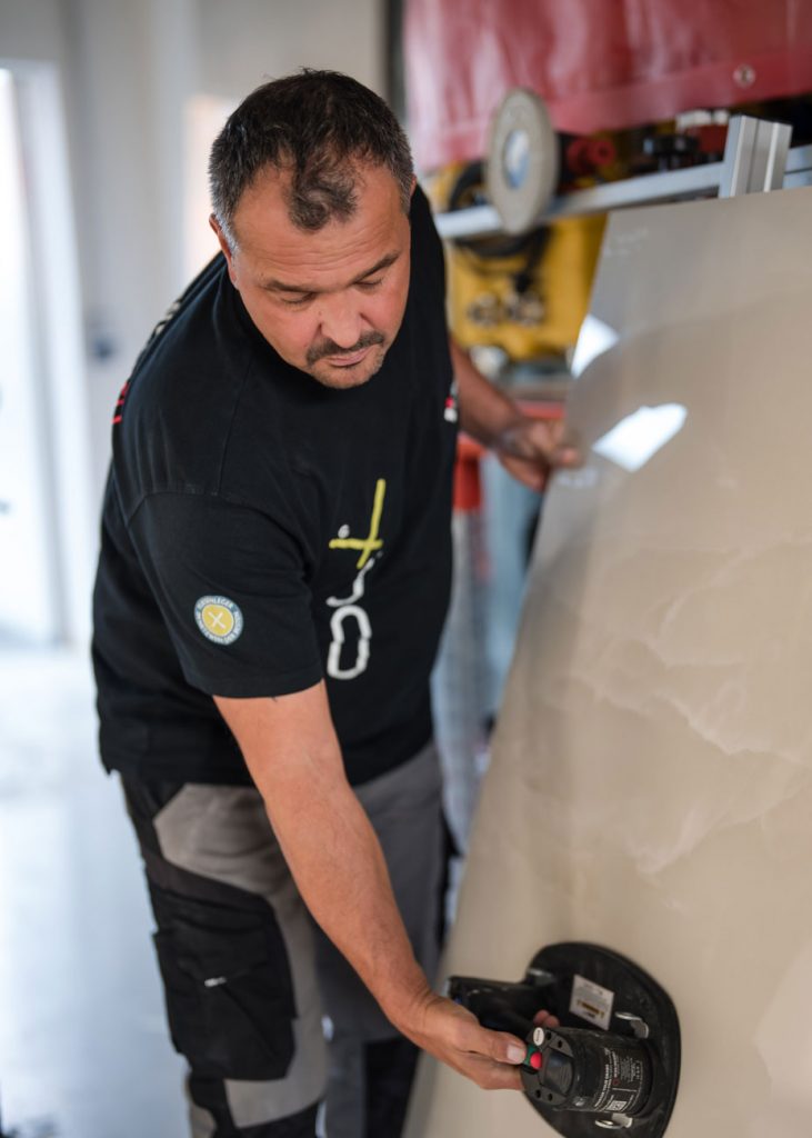 Ein zielstrebiger Fliegenleger in einem schwarzen T-Shirt und einer grauen Hose verwendet ein Saugwerkzeug, um eine große, helle Fliese oder Platte in einem Innenraum anzuheben oder anzupassen.