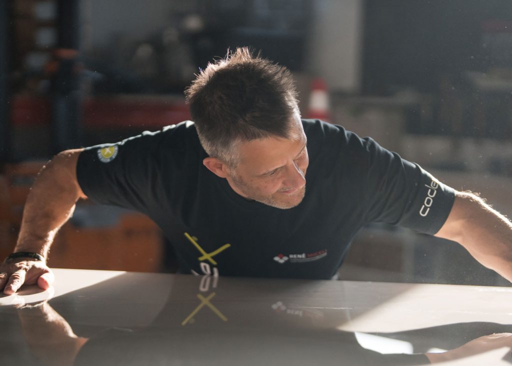 Ein Mann mit kurzen Haaren und schwarzem Hemd beugt sich vor und prüft sorgfältig eine glänzende weiße Oberfläche in einer Werkstatt. Das Sonnenlicht beleuchtet sein Gesicht und seine Arme - vielleicht ein engagierter Fliegenleger bei der Arbeit in Karlsruhe.