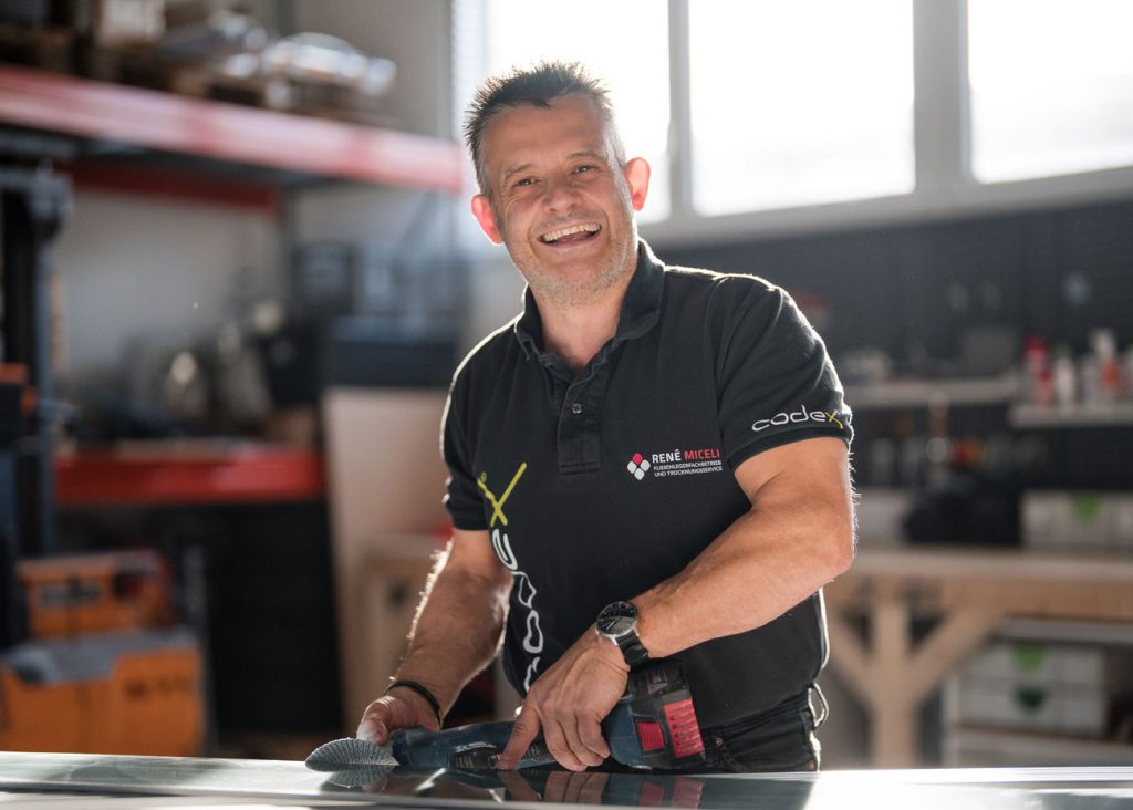 Ein lächelnder Mann in einem schwarzen Polohemd arbeitet mit Werkzeugen in einer hell erleuchteten Werkstatt. Er steht hinter einer Glasfläche, auf der im Hintergrund Regale und Geräte für die Bautrocknung und -veredelung zu sehen sind.