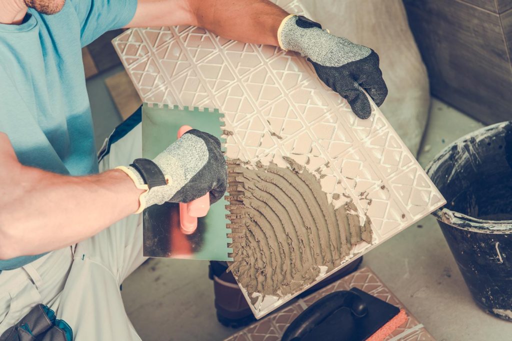 Ein Fliesenleger mit Handschuhen trägt mit einer Zahnkelle Kleber auf die Rückseite einer Dekorfliese auf, um sie für die Verlegung vorzubereiten. Ein Eimer mit mehr Klebstoff steht in der Nähe.