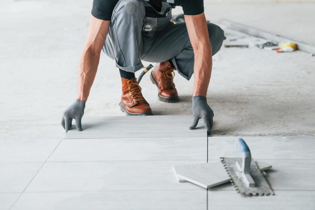 Ein Fliesenleger mit Handschuhen, Arbeitsstiefeln und grauer Hose kniet und verlegt sorgfältig große graue Bodenfliesen auf einer ebenen Fläche. In der Nähe sind Werkzeuge und weitere Fliesen zu sehen, die die präzise Arbeit der Fliesenverlegung zeigen.