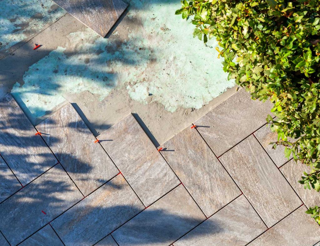 Diagonale Steinfliesen werden von einem erfahrenen Fliesenleger auf einer Terrasse verlegt, mit orangefarbenen Abstandshaltern zwischen einigen Fliesen. Ein Teil der Fliesenverlegung ist unvollendet, und am Rand der Terrasse sind grüne Sträucher zu sehen.