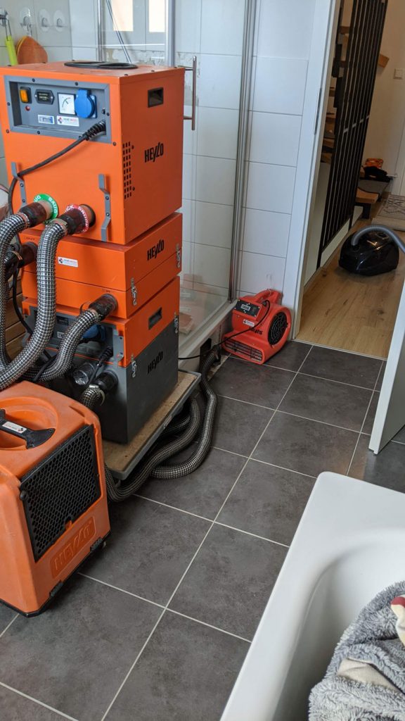 Mehrere orangefarbene Industrietrockner für Bautrocknung sind in einem gefliesten Badezimmer aufgestellt, wahrscheinlich nach einem Wasserschaden. Die Maschinen sind gestapelt oder auf dem Boden aufgestellt, und Schläuche verlaufen in einen angrenzenden Raum, was die Fliesenverlegung und die estriche Trocknung unterstützt.