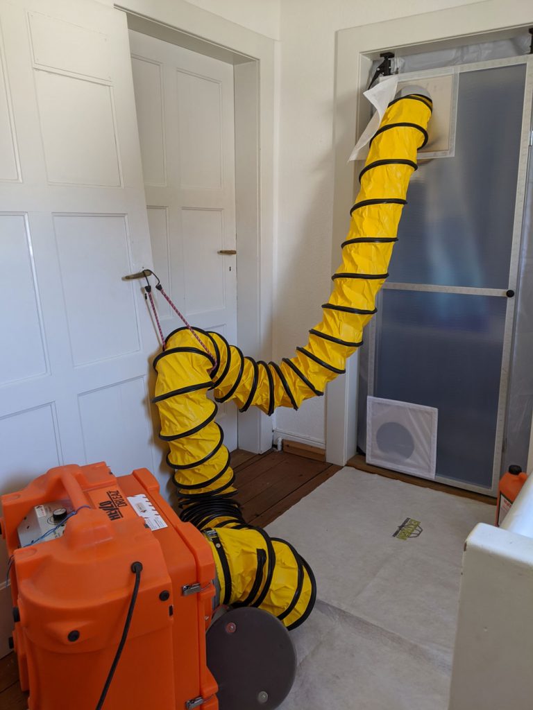 Eine große orangefarbene Maschine ist an einen flexiblen gelben Belüftungsschlauch angeschlossen, der in eine abgedichtete Türöffnung führt, die möglicherweise zur Bautrocknung oder Eindämmung durch einen Fliegenleger während Bau- oder Sanierungsarbeiten in einem Haus in Stutensee eingerichtet wurde.