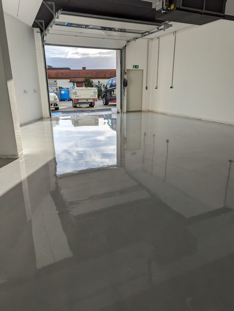 Eine saubere, leere Garage in Stutensee mit glänzendem, frisch beschichtetem grauen Epoxidboden über glattem Estrich. Das Garagentor ist offen und gibt den Blick frei auf Fahrzeuge und Häuser unter einem teilweise bewölkten Himmel.