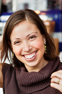 Eine Frau mit glattem braunem Haar und einem breiten, freundlichen Lächeln trägt einen braunen Pullover und Ohrringe. Sie steht in einem Haus und blickt in die Kamera, wobei sie mit einer Hand ihr Haar berührt. Der Hintergrund ist unscharf.