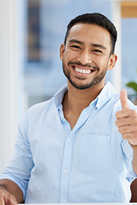 Ein lächelnder Mann mit kurzen dunklen Haaren und gestutztem Bart zeigt den Daumen nach oben. Er trägt ein hellblaues Button-down-Hemd und sitzt in einem Haus mit unscharfem Hintergrund.