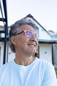 Ein Mann mittleren Alters mit grauem Haar und Brille steht lächelnd im Freien vor einem modernen weißen Haus mit großen Fenstern und einem spitzen Dach. Er trägt ein hellblaues Hemd.